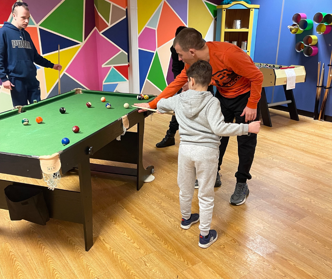 Two adults and a child are playing pool. One adult is helping the child to hit the ball with a snooker que.