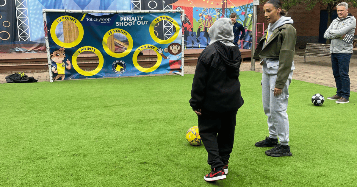 Two people are standing at a pop up football pitch with the young person about to kick a football into some pop up goals.